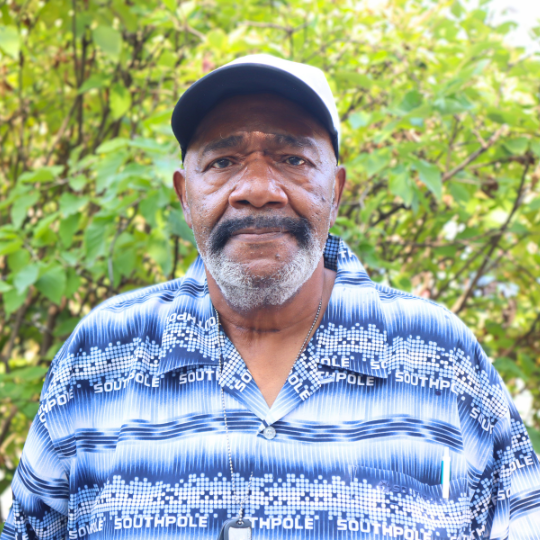 A headshot of a CAC member, Mr. Shanks, Sr. Mr Shanks, Sr. is wearing a white baseball cap and a collared shirt with a blue and white design. He is standing in front of a bush with green leaves.