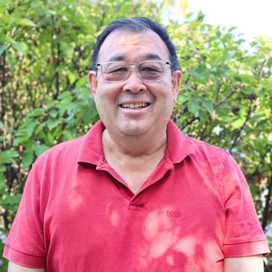 A photo of CAC member, Ron, smiling. Ron is wearing eyeglasses and a red button-up shirt, and is standing in front of a bush with green leaves.