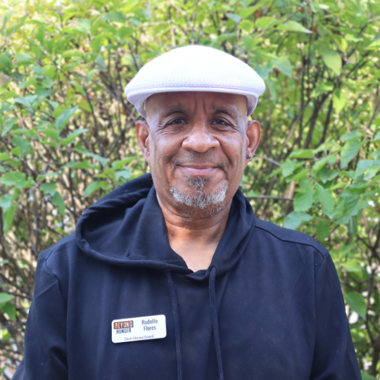 A headshot of a CAC member, Rudy. Rudy is wearing a white hat and black hooded sweatshirt, and is standing in front of a bush with green leaves.