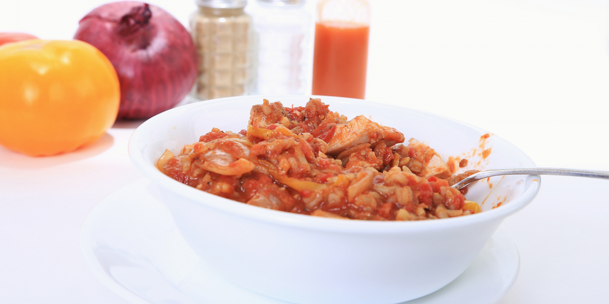 A white bowl of Chicken Creole, against a white background. Various items such as salt and pepper are in the background of the photo.